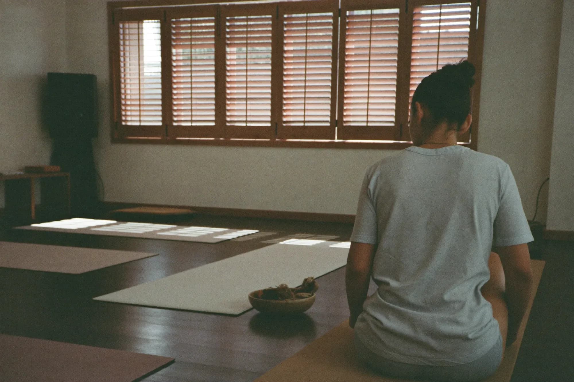 An instructor sitting quietly in an empty studio before class, mats laid out and light coming through the shutters.