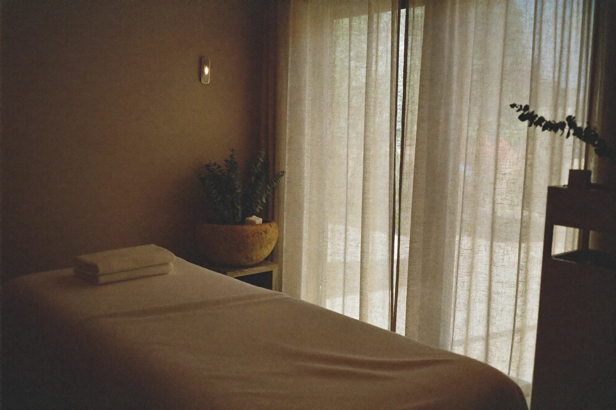 A spa treatment room with a massage table, folded towels and linen curtains drawn against the afternoon light.